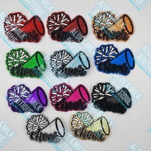 May include: A collection of embroidered patches featuring a megaphone and pom-pom design. Each patch is a different color, including red, orange, green, blue, purple, pink, black, silver, and gold. The word "cheer" is written in a cursive font on each patch.