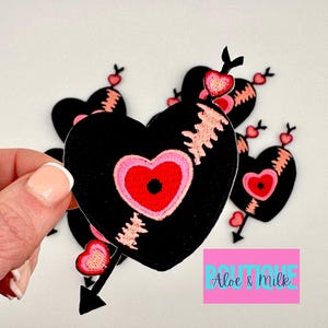 May include: A set of five black felt heart-shaped patches with pink and red stitching. Each heart has a pink and red heart inside with a red circle in the center. Each heart is pierced by a black arrow with a pink heart on the end.