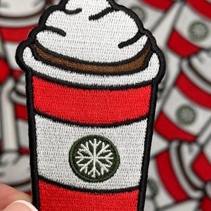 May include: Embroidered patch featuring a coffee cup design. The cup has a red base, white middle section, and a white whipped cream top with a brown stripe. A green snowflake emblem is on the cup.