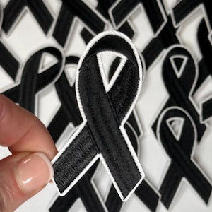 May include: A close-up of a black ribbon with white stitching. The ribbon is in the shape of a bow and is being held by a hand. The ribbon is surrounded by other black ribbons.