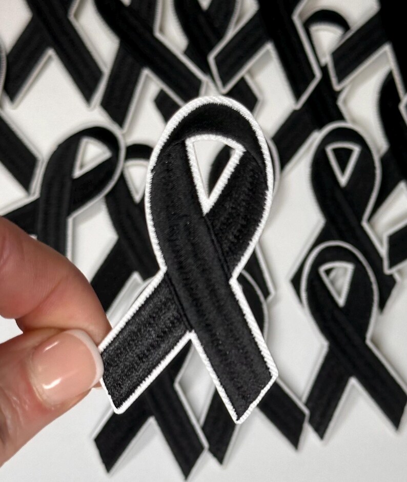 May include: A close-up of a black ribbon with white stitching. The ribbon is in the shape of a bow and is being held by a hand. The ribbon is surrounded by other black ribbons.