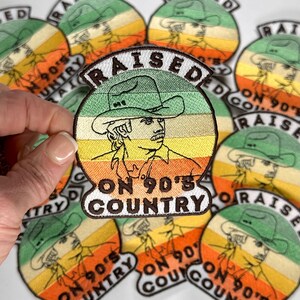 May include: A collection of embroidered iron-on patches with a country music theme. The patches feature a retro-style design with a cowboy wearing a hat and the text "Raised on 90's Country".