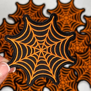 May include: Black and orange spiderweb patches. The patches feature a black background with an orange spiderweb design. The edges of the patches are also black. The patches are likely for Halloween or spooky-themed crafts.