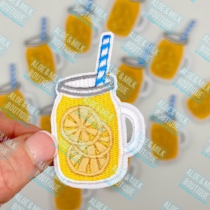 May include: A yellow embroidered iron-on patch of a mason jar with a blue and white striped straw and lemon slices inside. The patch is surrounded by other similar patches with the text "Aloe & Milk Boutique" repeated on each.