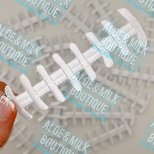 May include: White, embroidered football-shaped applique. The applique has a series of horizontal bars connected by a central vertical line. The text "ALOE & MILK BOUTIQUE" is visible in the background. This item is likely for crafting or embellishment.
