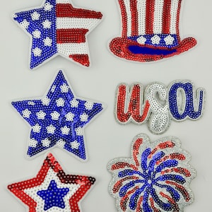 May include: A collection of six patriotic sequin patches featuring a star with a flag pattern, a top hat, a solid blue star with white dots, the text USA in script, a star with concentric red and white rings, and a firework burst design.
