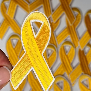 May include: A close-up of a yellow ribbon patch with white stitching. The patch is made of fabric and has a textured surface. The patch is in focus, and the background is blurred.
