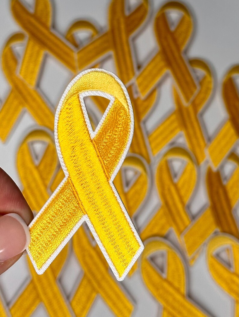May include: A close-up of a yellow ribbon patch with white stitching. The patch is made of fabric and has a textured surface. The patch is in focus, and the background is blurred.