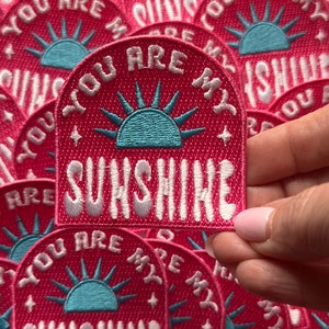 May include: A close-up of a pink fabric patch with a white embroidered sun and the text "You are my sunshine".