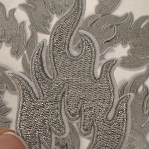 May include: A close-up of a silver embroidered flame patch. The patch is surrounded by other similar patches, all in various shades of gray.