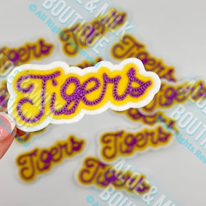 May include: A collection of yellow and purple "Tigers" patches. The word "Tigers" is embroidered in purple thread on a yellow background, outlined in white. The patches are scattered across a light background.