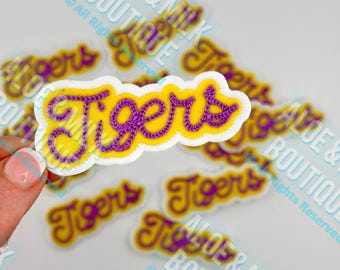 May include: A collection of yellow and purple "Tigers" patches. The word "Tigers" is embroidered in purple thread on a yellow background, outlined in white. The patches are scattered across a light background.