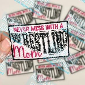 May include: Embroidered patch with the text "NEVER MESS WITH A WRESTLING Mom" in black and pink. The patch has a white background with a black border. Several patches are visible.