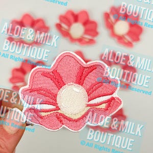 May include: Pink embroidered flower patch with a white pearl center. The patch is surrounded by white stitching. The patch is labeled "Aloe & Milk Boutique" in a blue font.