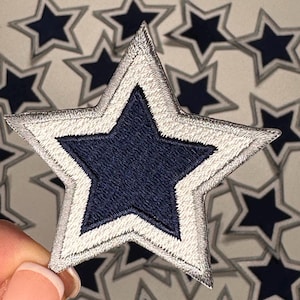 May include: A silver and navy blue star-shaped patch with a double outline. The patch is made of fabric and has a stitched design.