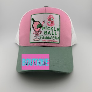 May include: A pink and green trucker hat with a white mesh back. The front of the hat features an embroidered patch with a pickleball cocktail and the text "Pickle Ball Cocktail Club Drink Responsibly!"