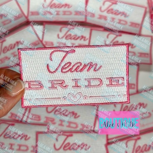 May include: White iron-on patch with pink embroidered text that reads "Team Bride". The patch has a pink border and a small pink heart in the center.