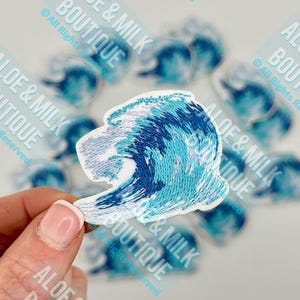 May include: A white embroidered patch with a blue and turquoise wave design. The patch is perfect for adding a nautical touch to clothing, bags, or other accessories.