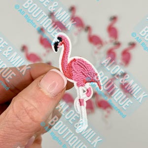 May include: A pink embroidered flamingo patch with a black beak and white legs. The patch is held in a hand, with other flamingo patches in the background. The text "ALOE & MILK BOUTIQUE" is visible.