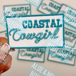 May include: A rectangular patch with the words "COASTAL Cowgirl" embroidered in teal thread on a white background. The patch has a teal border and is surrounded by other patches with the same design.
