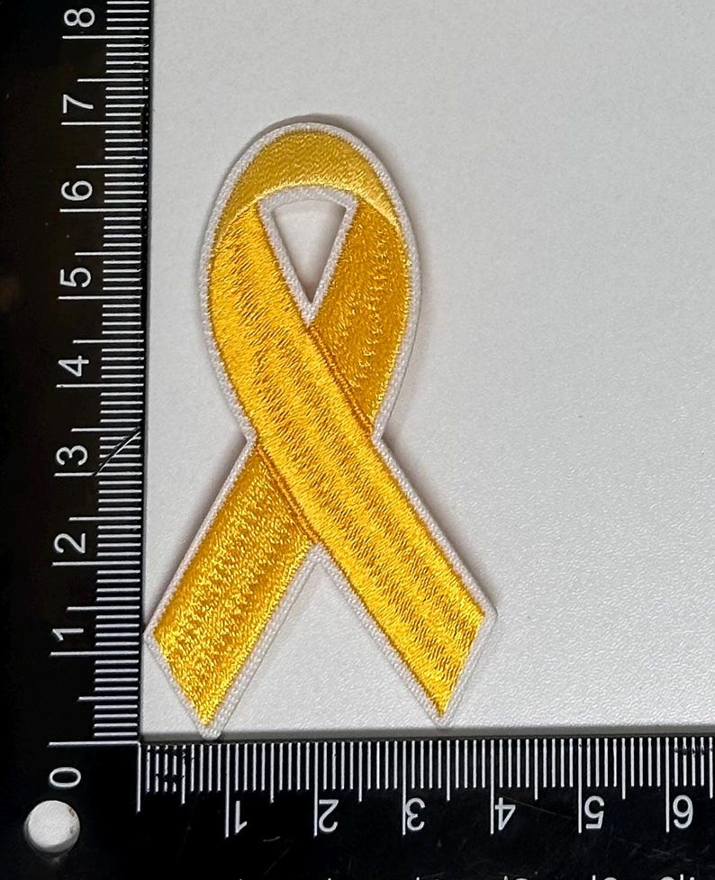 May include: A yellow ribbon embroidered patch with white stitching. The ribbon is tied in a bow shape.
