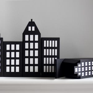 May include: Black and white cityscape decor featuring several building silhouettes with illuminated windows. The set includes various building shapes and sizes, creating a miniature urban scene. The decor is designed to stand on a shelf or tabletop.
