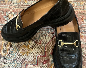 May include: A pair of black leather loafers with gold buckles. The loafers have a crocodile-embossed pattern and a chunky platform sole.