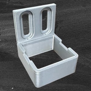 Op de afbeelding: Een zilverkleurige, 3D-geprinte beugel met een rechthoekige basis en twee verticale, langwerpige openingen. De beugel heeft een getextureerd oppervlak en is ontworpen om een object vast te houden. De achtergrond is donkergrijs.