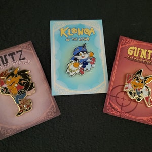 May include: Three enamel pins featuring characters from video games. The first pin is a red, yellow, and black wolf character with the text "Butz Bounty Hunter". The second pin is a blue, yellow, and red character with the text "Klonoa of the Wind". The third pin is a red, yellow, and black fox character with the text "Guntz Golden Killer".