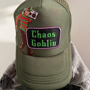 May include: Green trucker hat with a black and green embroidered patch that says "Chaos Goblin". The patch is above a cartoon image of a man holding a boombox.