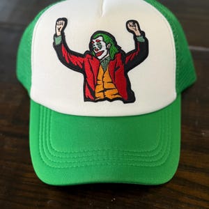 May include: Green and white trucker hat featuring an embroidered image of a character in a red suit, orange vest, and green hair. The character has white face paint and is raising their arms in a celebratory pose. The hat has a green brim and mesh back.