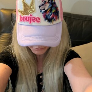 May include: A pink and white trucker hat with a gold crown and a colorful lion patch. The hat has the word "boujee" written on the front.