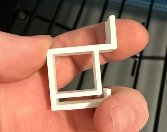 May include: A small, white, plastic bracket with a rectangular opening and a right-angle extension. The bracket is held in a hand, with a dark background of metal bars and a blue surface.