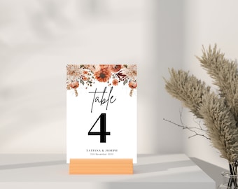 May include: A white table number card with a floral design in shades of orange, brown, and cream. The card reads "Table 4" and "Tatiana & Joseph 12th November XXXX".