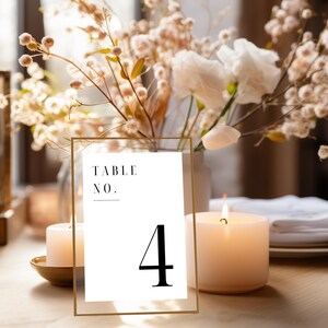 Può includere: Un biglietto bianco con testo nero che dice "Table No. 4" in una cornice dorata. Il biglietto è su un tavolo con una candela accesa e un vaso di fiori secchi.