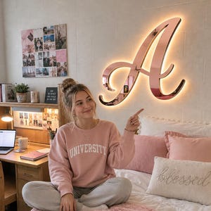 May include: A large, rose gold, script-style letter A wall sign with integrated backlighting mounted on a white wall. The sign features a reflective surface and emits a warm glow, positioned above a bed with pink and white bedding and pillows.