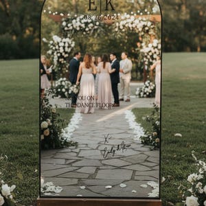 May include: A large, arched mirror with a wooden base, displaying wedding details. The top features initials "EK" and the date "08.26.2024." Text below reads "See you on the dance floor" and includes a signature. The scene reflects a wedding ceremony.
