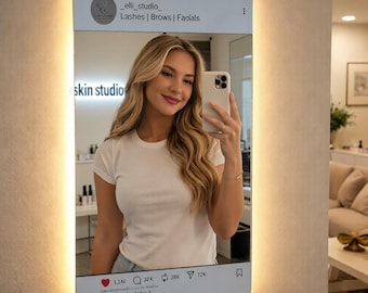 May include: A decorative mirror shaped like a smartphone, with a woman taking a selfie. The mirror has a glowing border and features social media-style icons and text, including "_elli_studio_ Lashes | Brows | Facials" and "skin studio".
