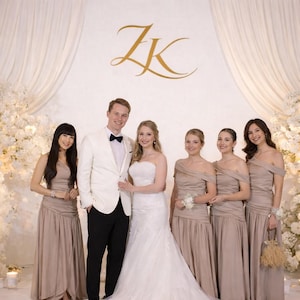 May include: A wedding group poses in front of a white floral backdrop. The bride wears a white strapless gown, and the groom wears a white suit with a black bow tie. Bridesmaids wear taupe dresses. Gold initials are on the wall.