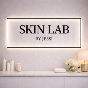May include: A rectangular illuminated sign with the words "SKIN LAB" in black, with "BY JESSI" underneath. The sign is framed in black and mounted on a white wall. Below, a white countertop displays white bottles, a lamp, and a floral arrangement.