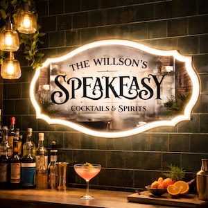 May include: An oval-shaped, backlit mirror sign with a decorative scalloped edge. The text reads THE WILLSON'S SPEAKEASY COCKTAILS & SPIRITS in black lettering. The mirror is mounted on a dark green tiled wall above a bar counter with glassware.