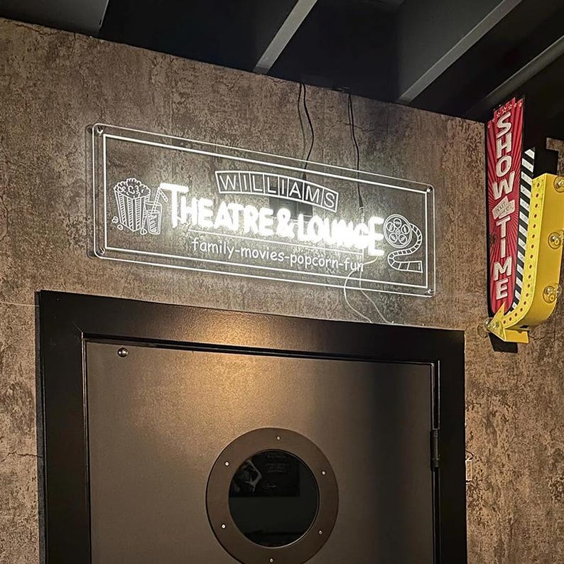 May include: A white neon sign with the text "Williams Theatre & Lounge" and the tagline "family-movies-popcorn-fun". The sign is mounted on a brown brick wall. A red and yellow "Showtime" sign is visible in the background.