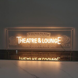May include: A white illuminated sign with the text "MININNO THEATRE & LOUNGE" and the words "movies-popcorn-drinks-games" below. The sign is lit from behind, creating a warm glow.