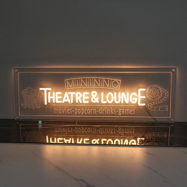 May include: A white illuminated sign with the text "MININNO THEATRE & LOUNGE" and the words "movies-popcorn-drinks-games" below. The sign is lit from behind, creating a warm glow.