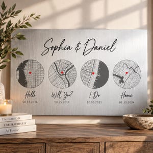 Pode incluir: Uma placa de parede retangular de metal escovado com os nomes Sophia & Daniel em letra cursiva preta. Abaixo, quatro ilustrações de mapas circulares, cada uma marcada com um pequeno coração vermelho e rotulada com datas e texto.