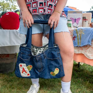 Op de afbeelding: Een blauwe denim tote bag met een witte aas van harten patch en groene olijftak borduurwerk. De tas wordt vastgehouden door een persoon die een rood-zwart geometrisch print shirt en denim shorts draagt.