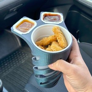 Może przedstawiać: Czarny plastikowy pojemnik na żywność z trzema przegródkami. Pojemnik ma wbudowany uchwyt i zawiera nuggetsy z kurczaka i dwa różne sosy do maczania.