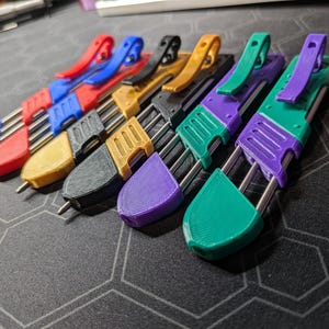 May include: A set of six multi-colored pocket knives with a clip on the top. The knives are arranged in a row from left to right, with the colors being blue, red, gold, black, purple, and green.