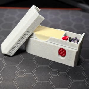 Può includere: Un portadadi rettangolare grigio chiaro con la scritta "SIDESTRIKE" sul coperchio. La scatola è aperta, rivelando una carta gialla, un oggetto rosso e diversi dadi a più facce. Un disegno circolare rosso è sulla parte anteriore della scatola.