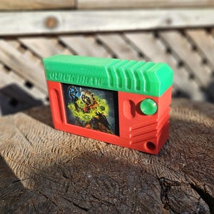 May include: A red and green Quick Draw card holder. The top is green with ridges, and the front has a card with a colourful illustration. A green button is on the side.